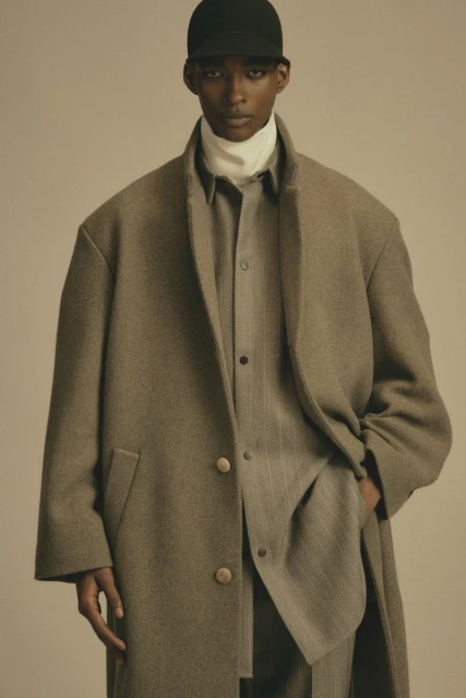 Fashion portrait of a young adult Black man in a wool coat, baseball cap, and suit in a neutral studio setting with soft lighting.