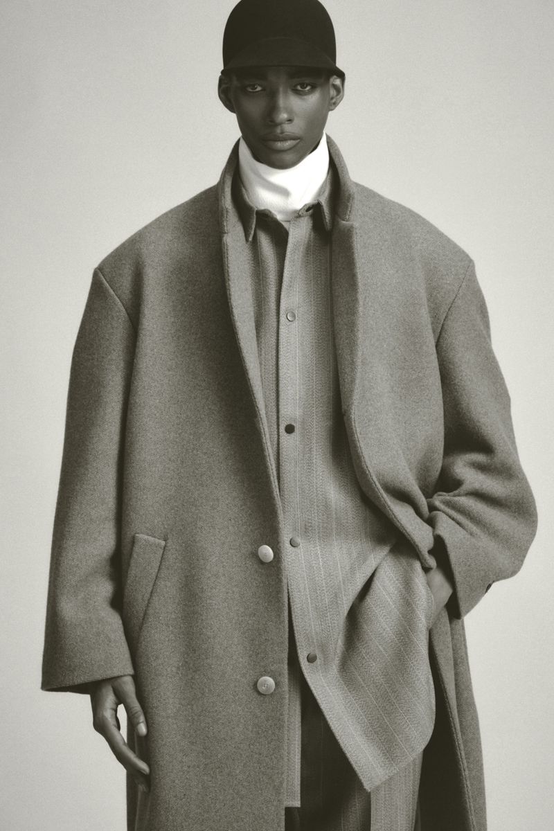 Monochrome portrait of a Black male model wearing a cap, turtleneck, and overcoat, against a neutral backdrop in a studio setting.
