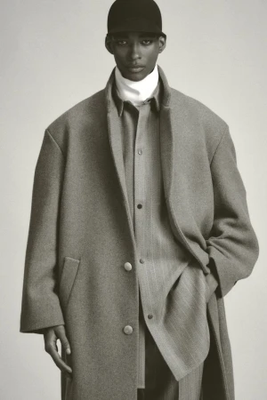 Monochrome portrait of a Black male model wearing a cap, turtleneck, and overcoat, against a neutral backdrop in a studio setting.