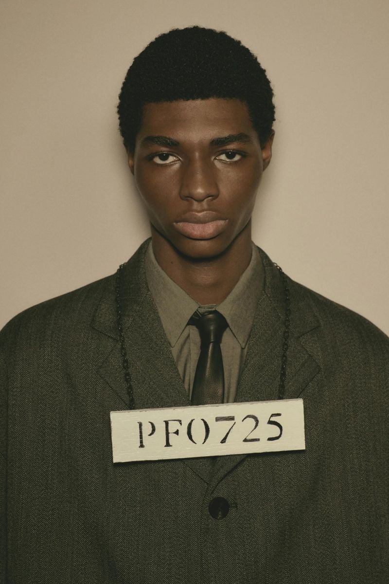Close-up portrait of a young adult Black man wearing a suit and tie, with a placard around his neck against a plain light background.