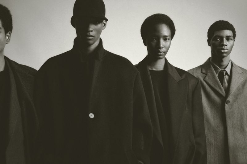 Four African American men stand in a row, wearing black and gray suits against a grey background, photographed in a studio setting.