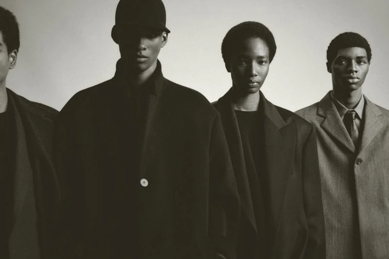 Four African American men stand in a row, wearing black and gray suits against a grey background, photographed in a studio setting.