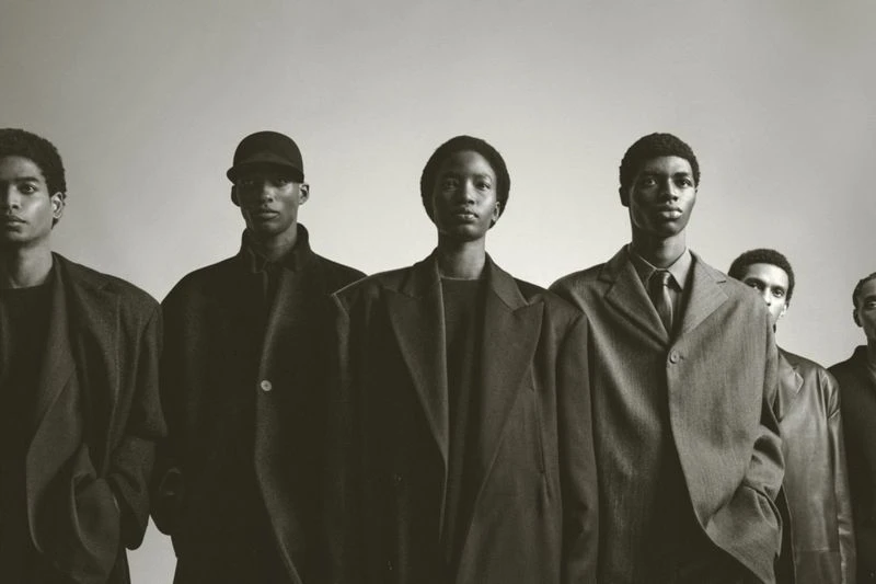 Monochrome fashion shot of a group of African American adult models in overcoats posing indoors in a photo studio, lit with soft lighting.