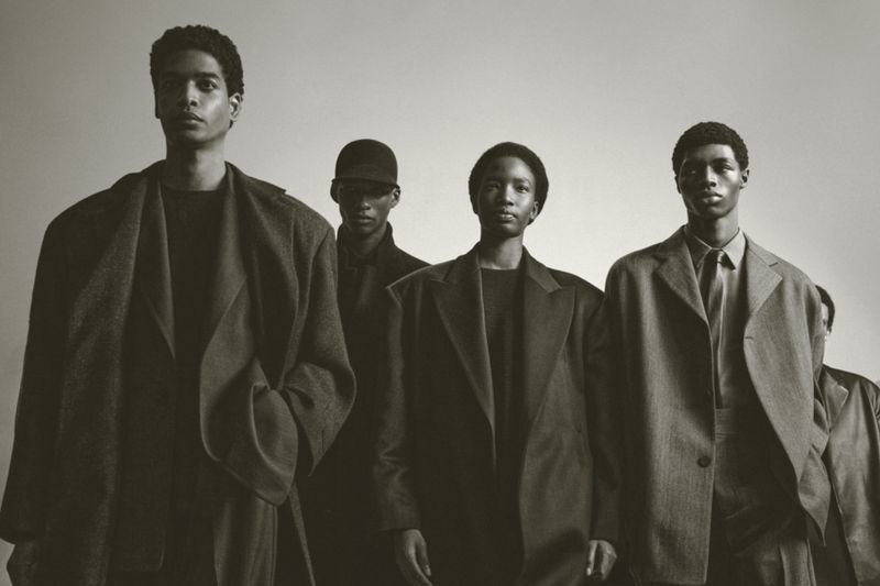 Five young adult males in a dimly lit indoor photo studio setting. They are wearing matching suits and coats in neutral hues.