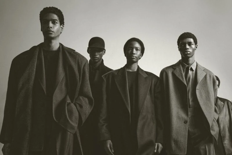 Five young adult males in a dimly lit indoor photo studio setting. They are wearing matching suits and coats in neutral hues.