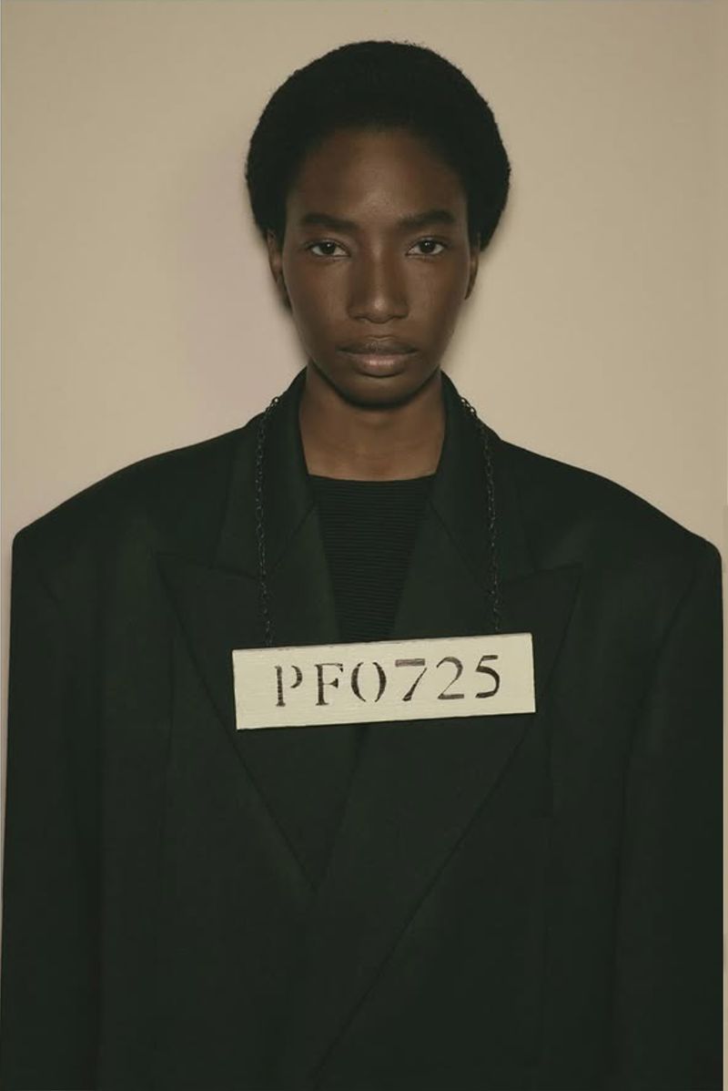 Close-up studio portrait of a Black female fashion model in a dark jacket wearing a numbered card hanging from a chain around her neck.