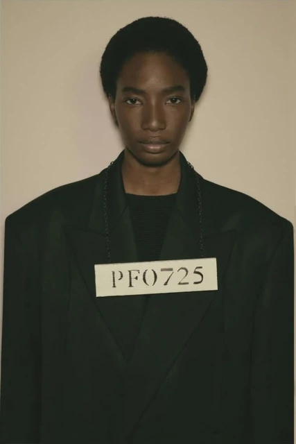 Close-up studio portrait of a Black female fashion model in a dark jacket wearing a numbered card hanging from a chain around her neck.