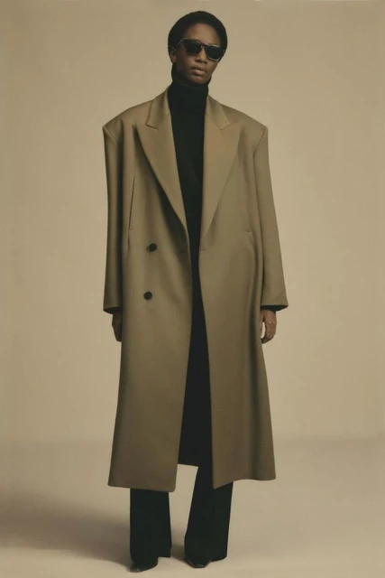 Adult model in oversized neutral coat and black turtleneck and pants against a plain background in a fashion photography studio shot.