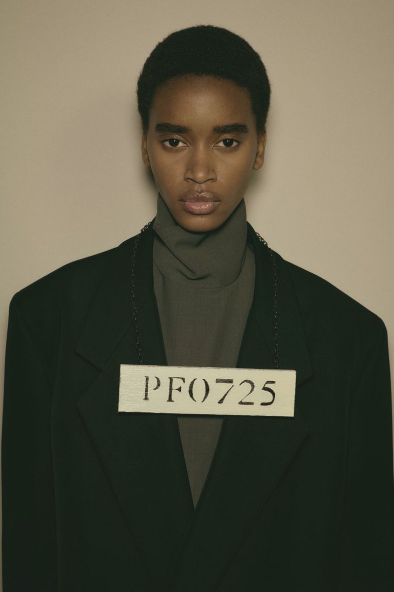Portrait of a black fashion model in a black blazer and grey turtleneck against a neutral studio backdrop, holding a number tag.