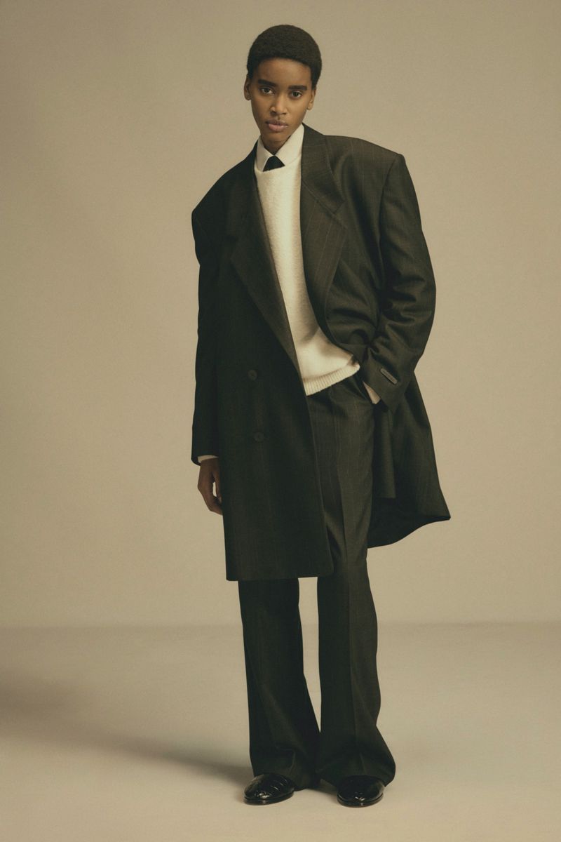 Model wearing a dark oversized coat over a white sweater against a plain neutral background