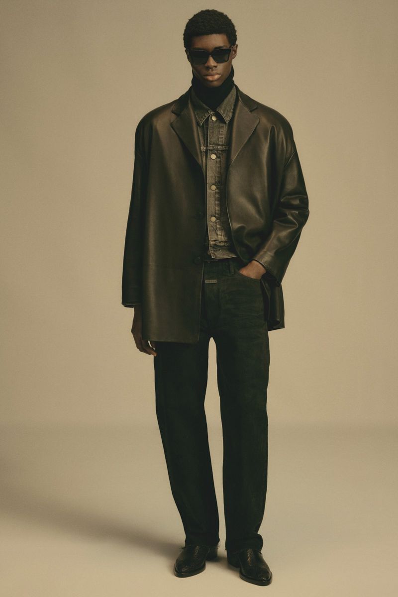 Adult man poses in studio wearing black leather jacket, black turtleneck, dark denim shirt, black slacks, and sunglasses in studio lighting.
