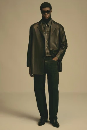 Adult man poses in studio wearing black leather jacket, black turtleneck, dark denim shirt, black slacks, and sunglasses in studio lighting.