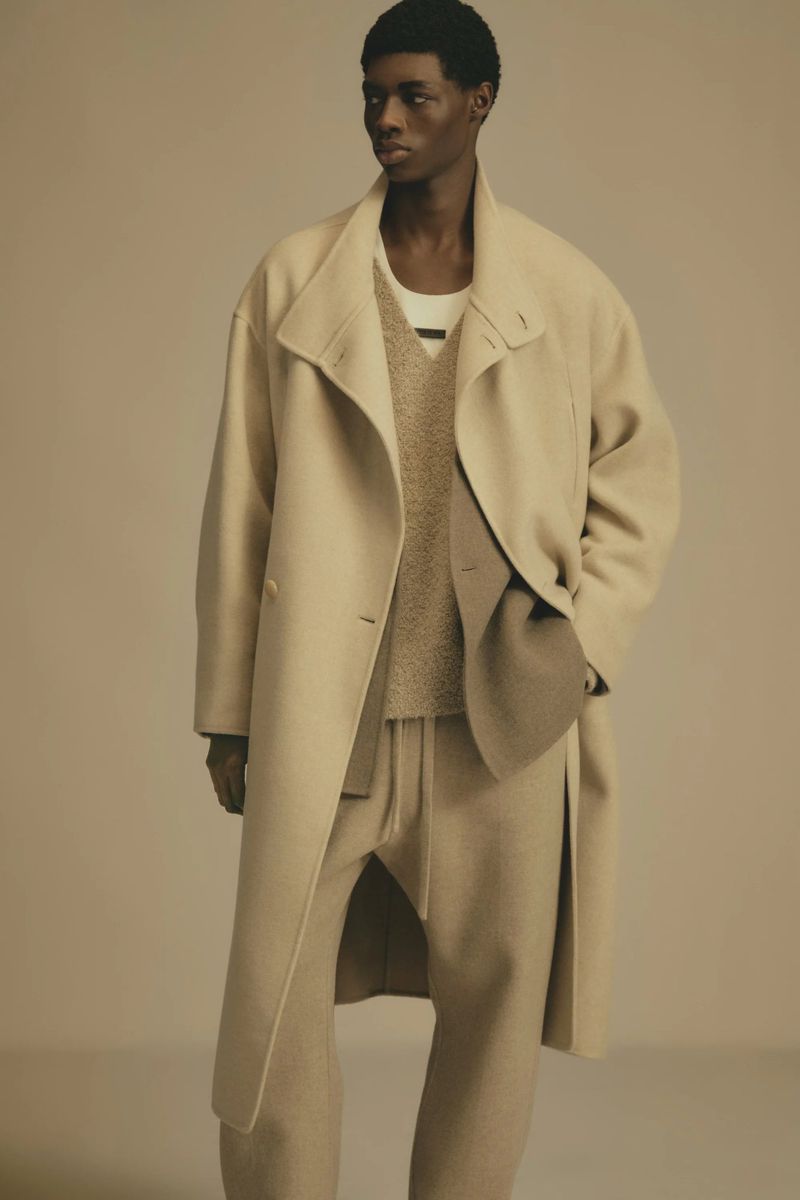 A male fashion model poses in a neutral studio setting wearing a beige overcoat, sweater, and loose-fitting trousers in a full-length shot.