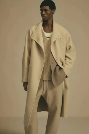 A male fashion model poses in a neutral studio setting wearing a beige overcoat, sweater, and loose-fitting trousers in a full-length shot.