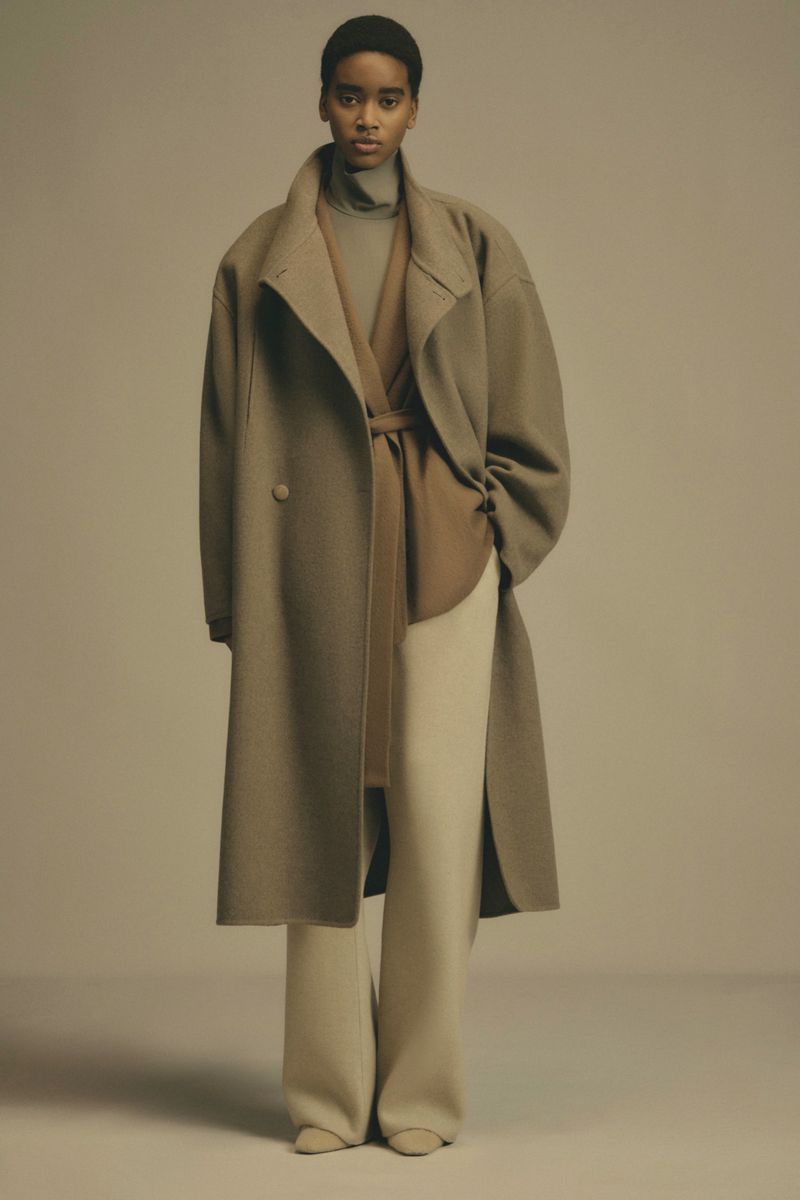 A woman models a beige, double-breasted overcoat with a belt, paired with a matching turtleneck and wide-leg pants in a neutral setting.