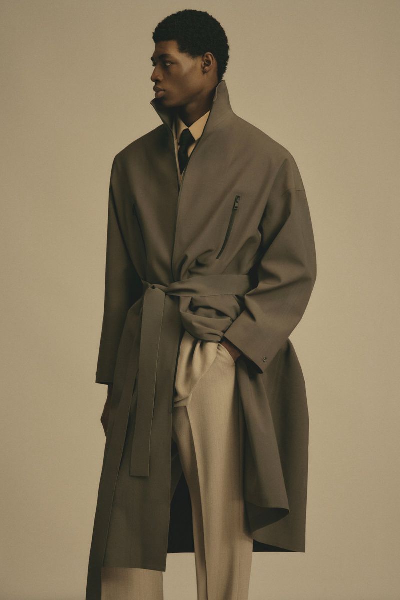 Studio shot of a Black male model wearing a belted overcoat and light pants, looking left, set against a neutral backdrop.