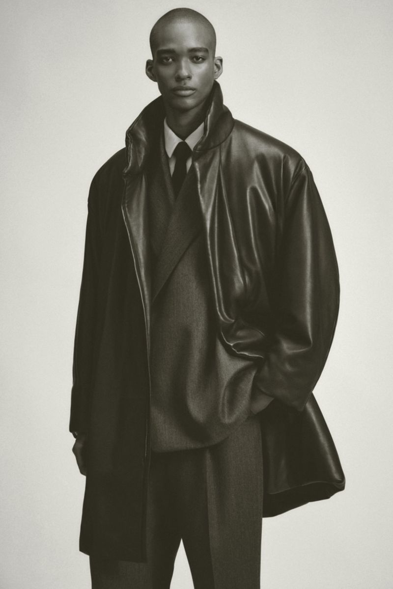 Model wearing a dark leather trench coat and suit against a plain light background