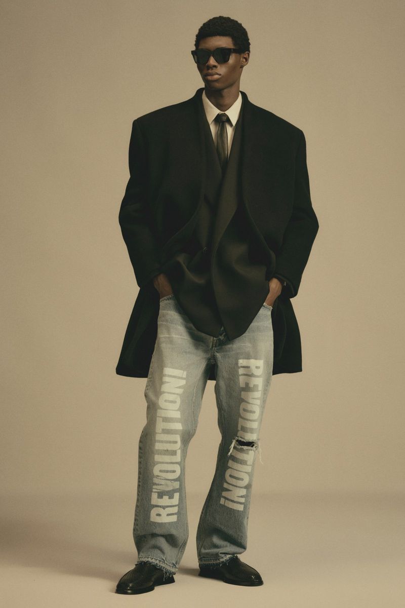 Man in black coat, vest, tie, light blue denim pants with white 'REVOLUTION!' text, and sunglasses in studio, full-length portrait.