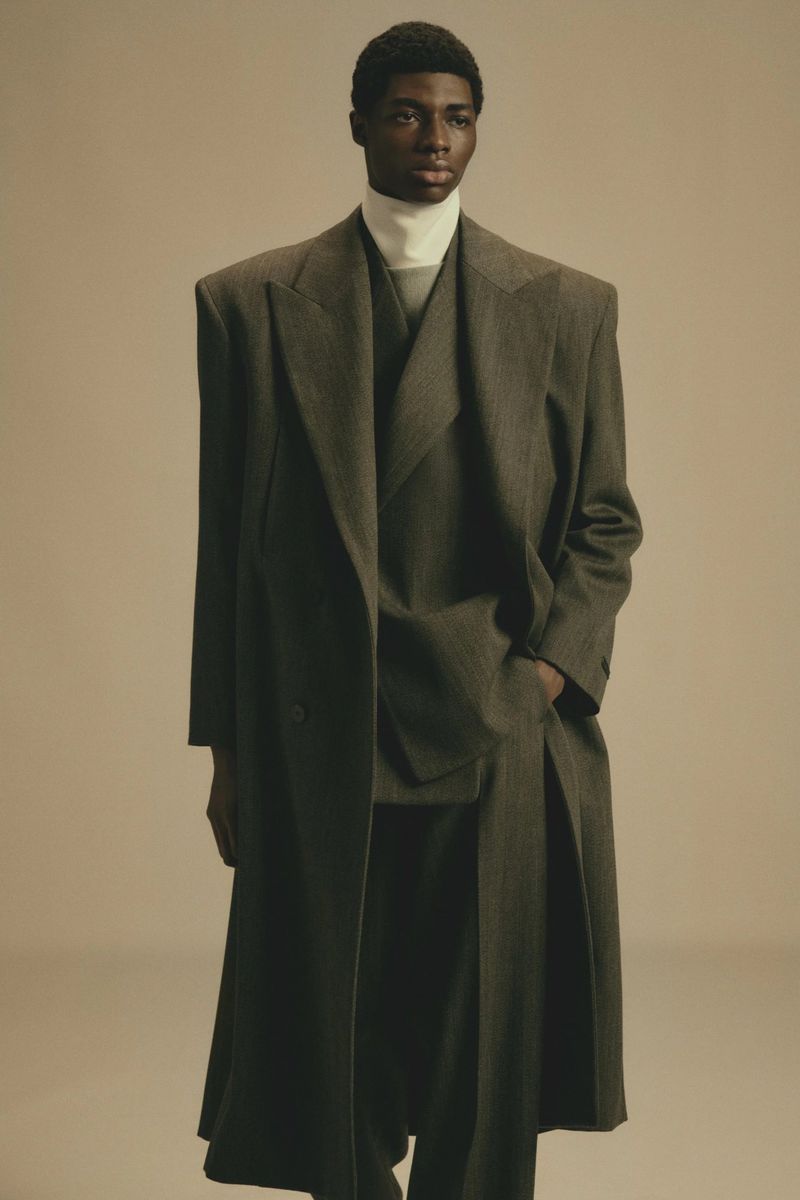 Full-length portrait of a dark-skinned adult male in a high-neck top, dark-green pantsuit, and matching overcoat against a light-beige background.