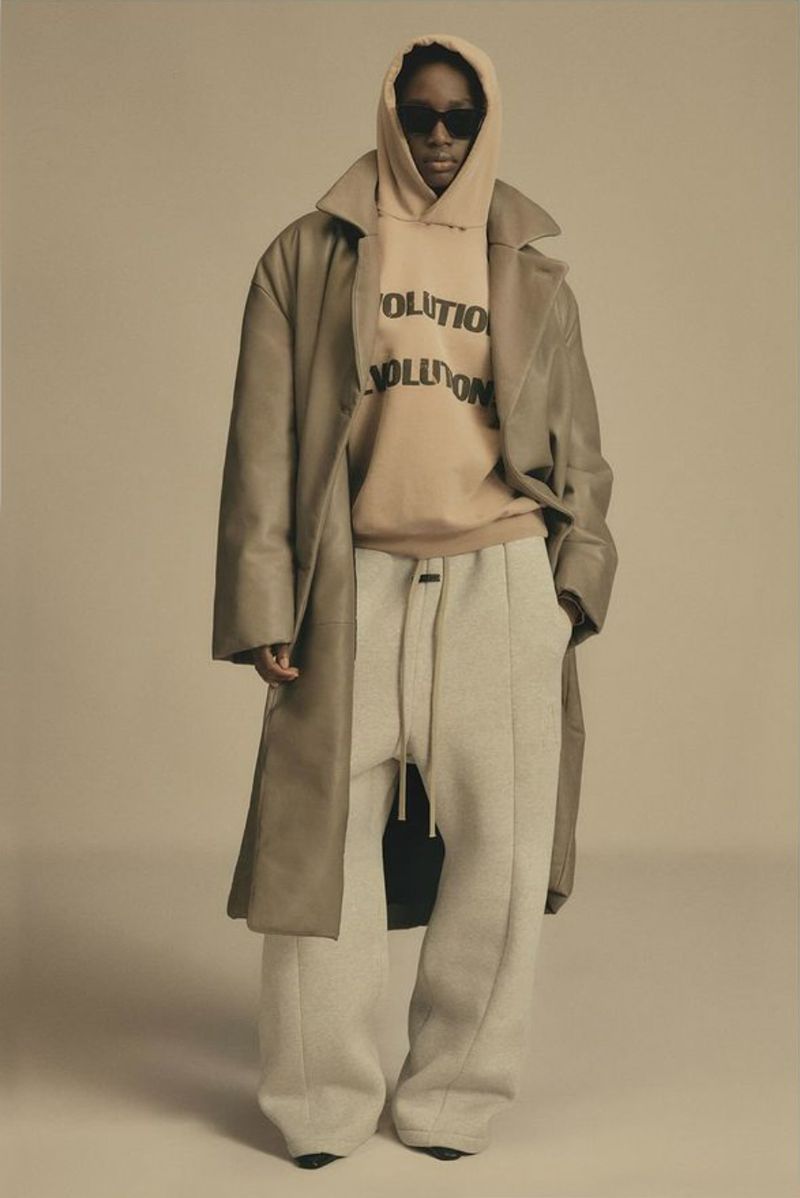 Person stands in studio wearing beige hoodie with 'REVOLUTION' text, light pants, and duster coat in a neutral fashion shot.