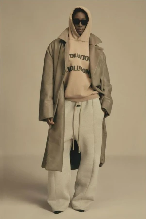 Person stands in studio wearing beige hoodie with 'REVOLUTION' text, light pants, and duster coat in a neutral fashion shot.