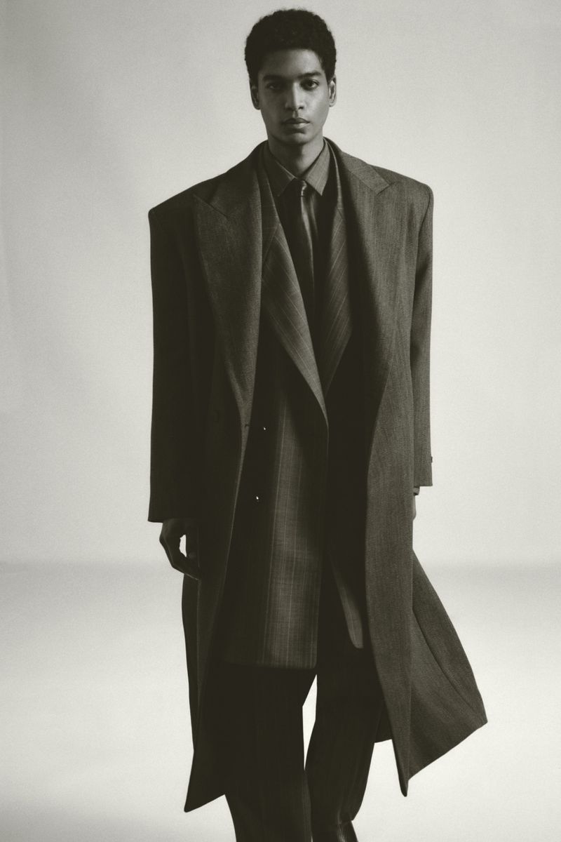 Fashion editorial shot of an African American man in a long, oversized coat, standing indoors against a neutral background.