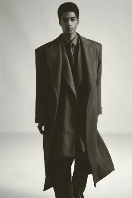 Fashion editorial shot of an African American man in a long, oversized coat, standing indoors against a neutral background.