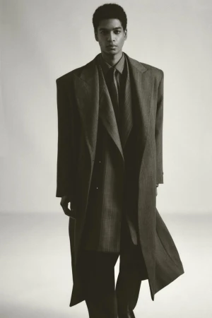 Fashion editorial shot of an African American man in a long, oversized coat, standing indoors against a neutral background.