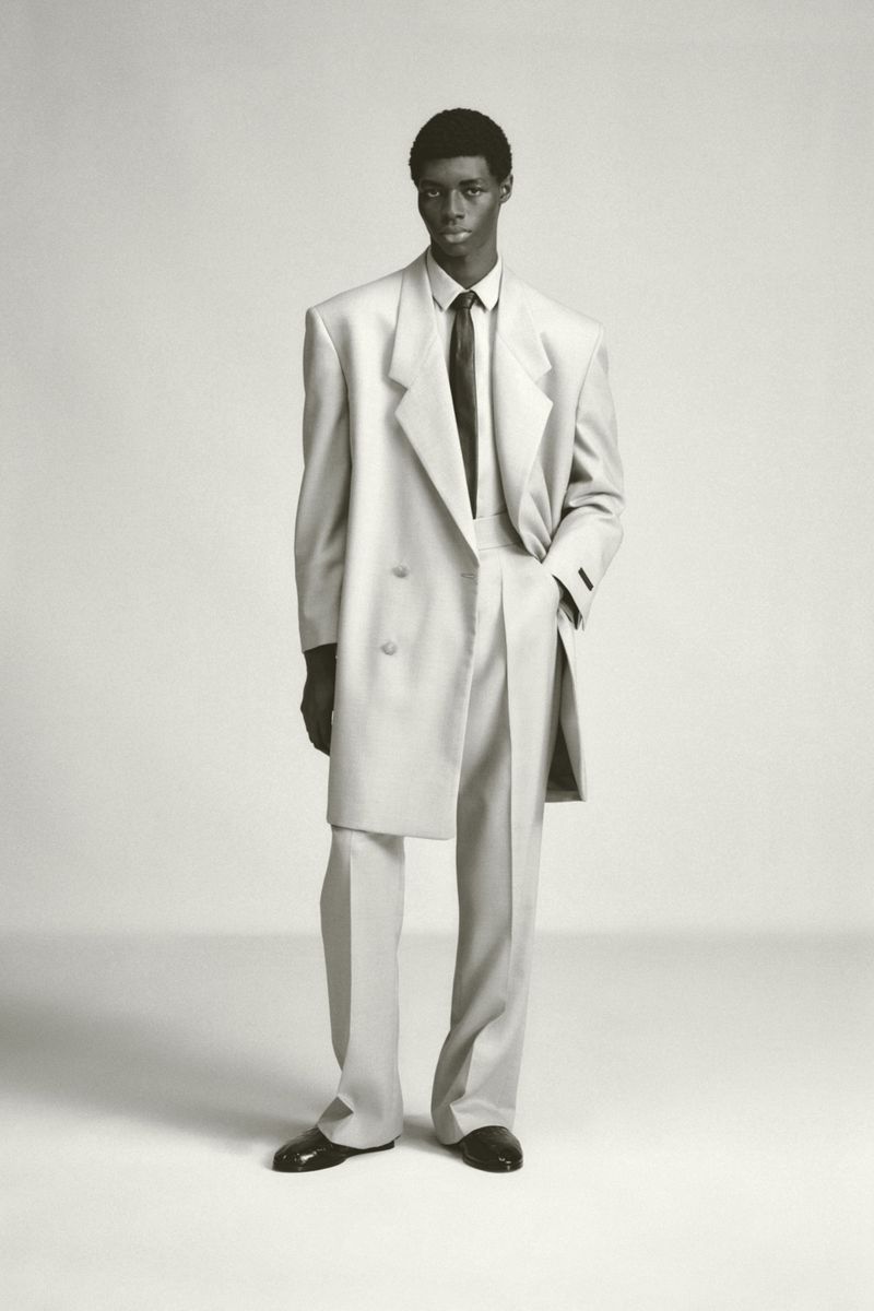 Monochrome studio shot of a Black male model in an oversized, light-colored double-breasted suit, black tie, and patent leather dress shoes.