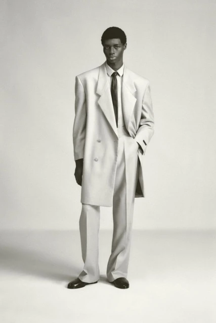 Monochrome studio shot of a Black male model in an oversized, light-colored double-breasted suit, black tie, and patent leather dress shoes.
