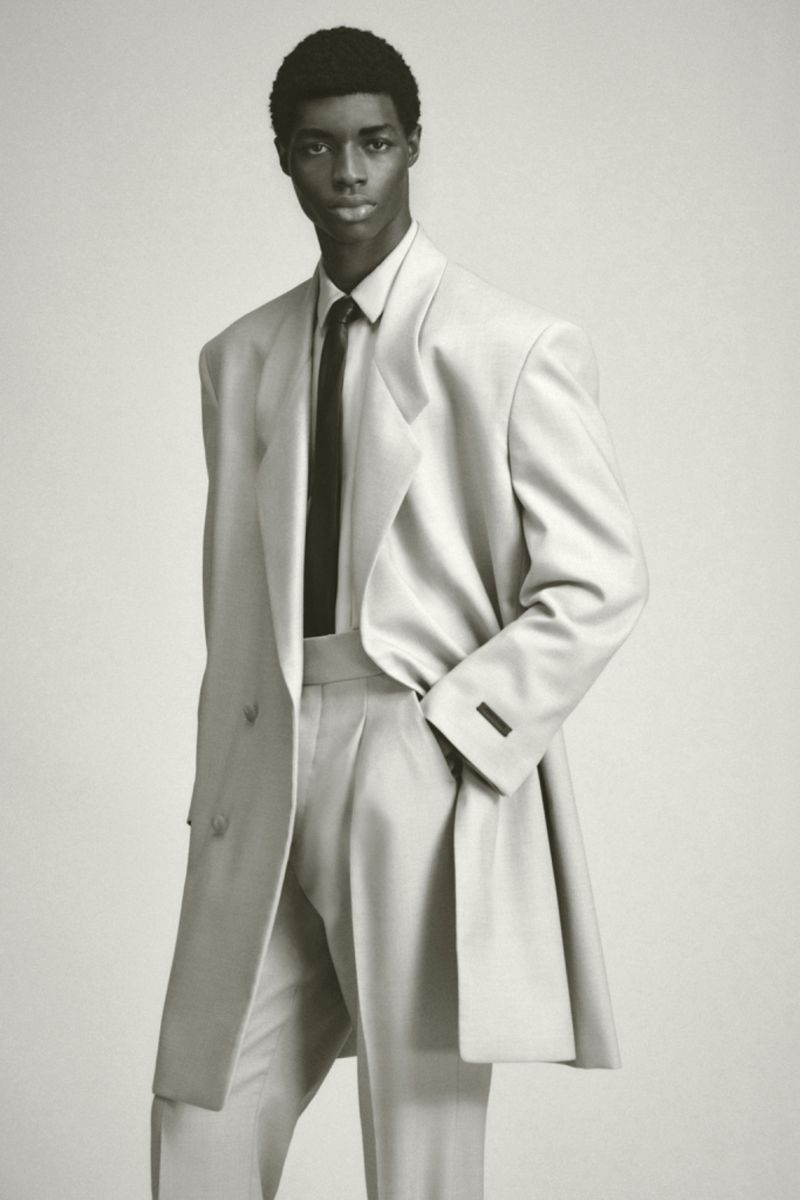 Man wearing a cream tailored coat, matching trousers, tie and shirt against a solid light background.