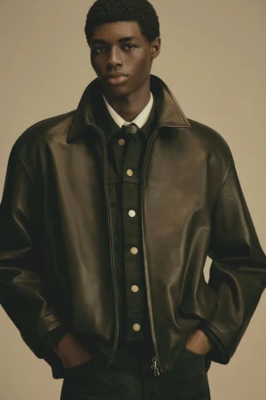 Portrait of an African American male model wearing a dark leather jacket with a buttoned shirt underneath, set against a neutral backdrop.