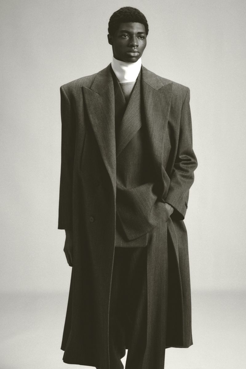 Studio shot of a Black male model in a grey double-breasted overcoat, matching suit, and a white turtleneck sweater, posing against a grey backdrop.