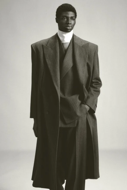 Studio shot of a Black male model in a grey double-breasted overcoat, matching suit, and a white turtleneck sweater, posing against a grey backdrop.