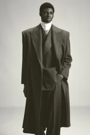 Studio shot of a Black male model in a grey double-breasted overcoat, matching suit, and a white turtleneck sweater, posing against a grey backdrop.