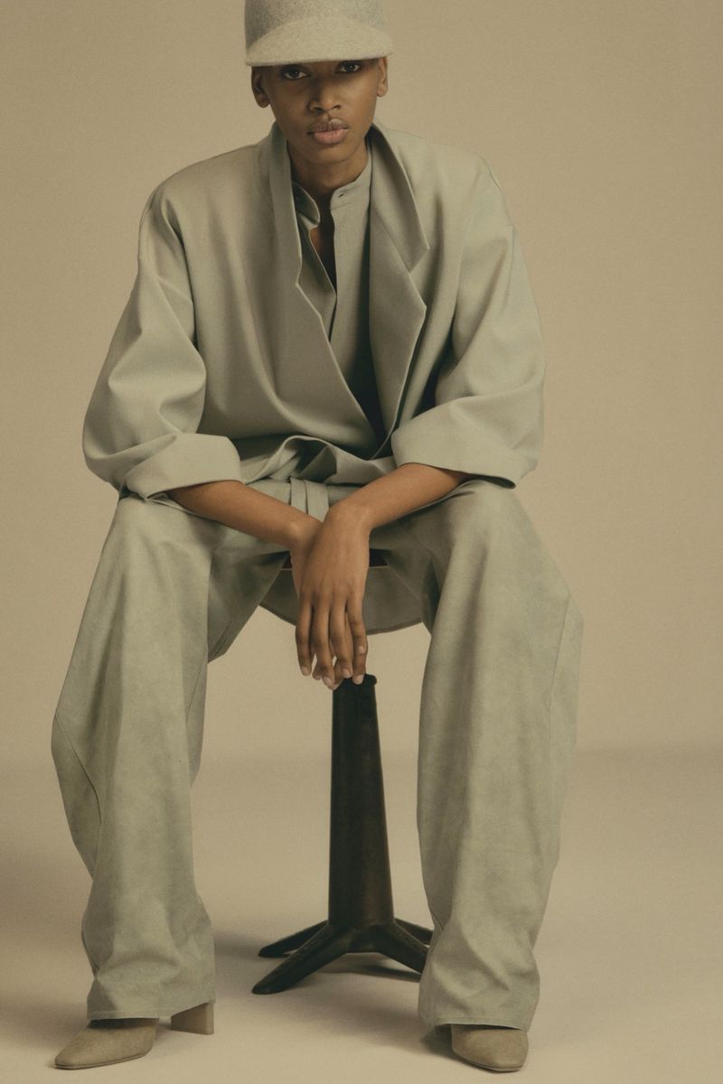 An adult woman is sitting on a stool wearing a long-sleeved, matching oversized pants and collared shirt and cap in a neutral-tone photo studio.