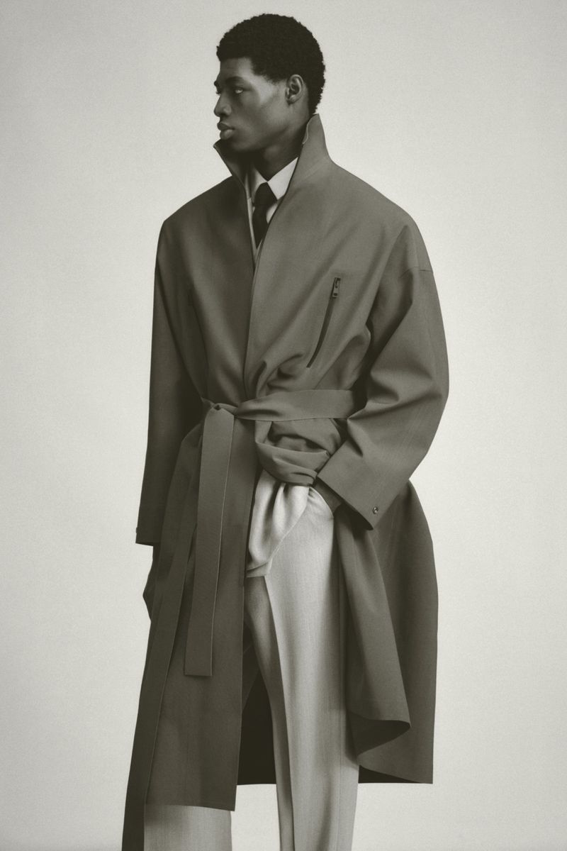 An African-American male model stands in a neutral-toned photo studio wearing an olive trench coat, light dress pants, and dark tie.