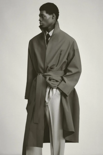 An African-American male model stands in a neutral-toned photo studio wearing an olive trench coat, light dress pants, and dark tie.