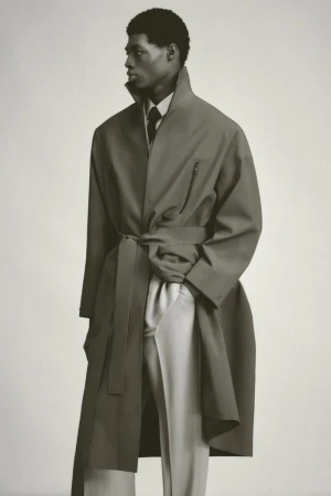 An African-American male model stands in a neutral-toned photo studio wearing an olive trench coat, light dress pants, and dark tie.