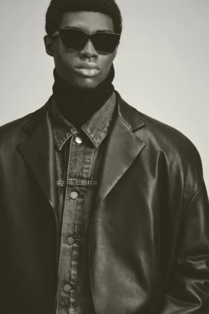Model in dark sunglasses wearing a black leather coat over a denim jacket against a neutral studio background