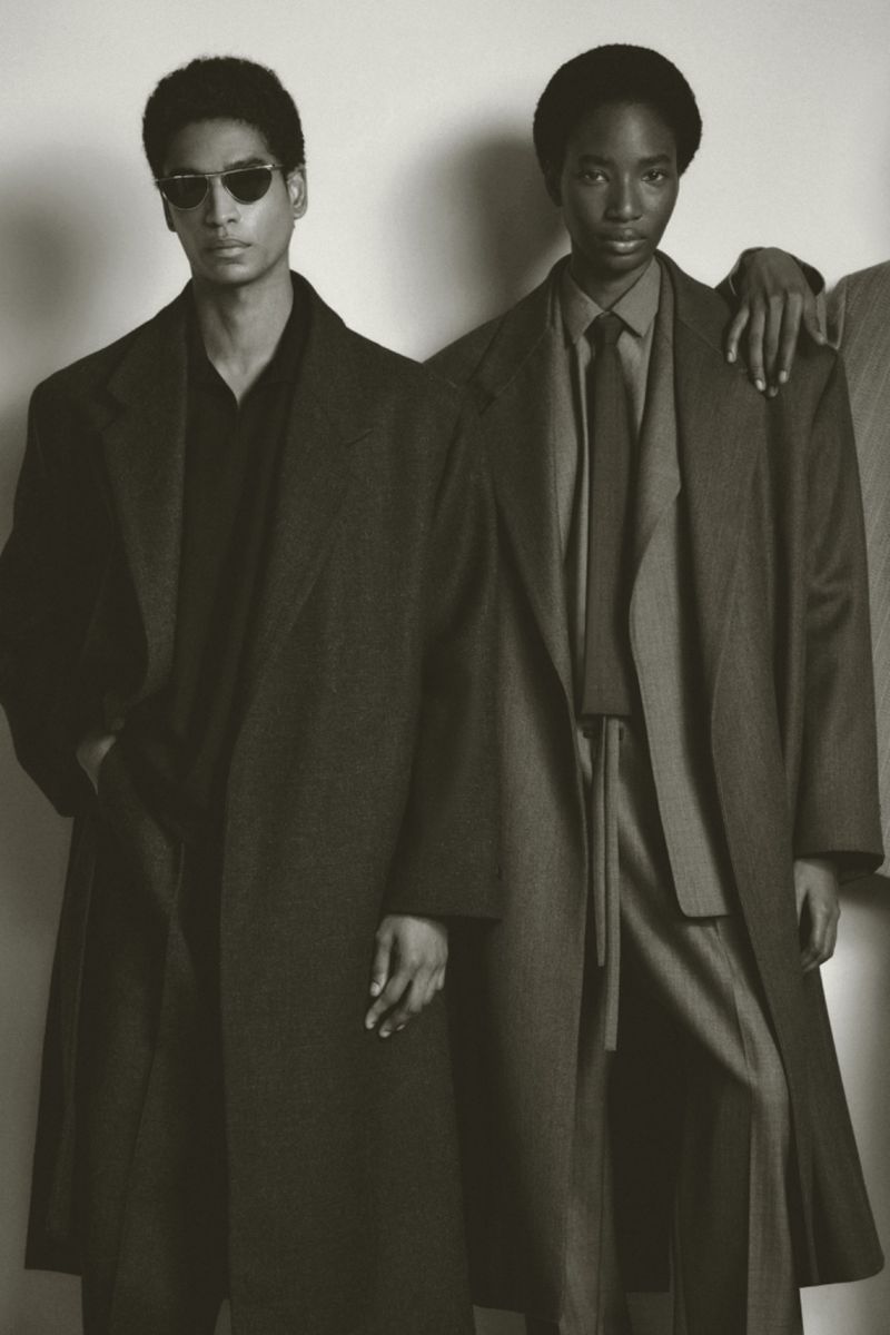 Two models wearing tailored oversized coats and sunglasses against a plain studio background.