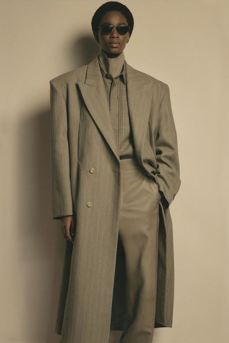 Woman in oversized pinstripe coat and tan pants, wearing dark sunglasses, standing against a neutral backdrop in a fashion editorial.