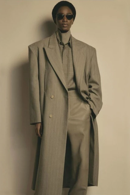 Woman in oversized pinstripe coat and tan pants, wearing dark sunglasses, standing against a neutral backdrop in a fashion editorial.