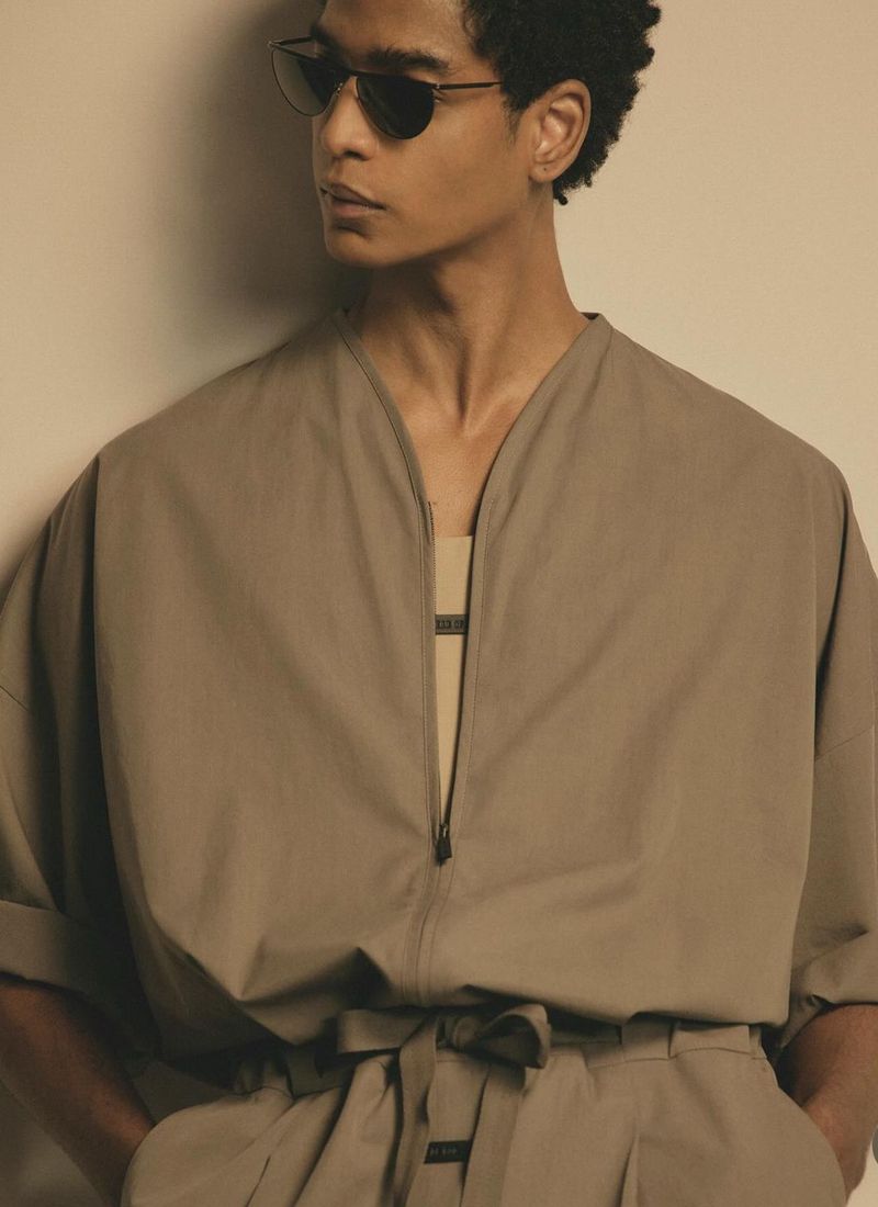 Man in stylish jumpsuit and sunglasses posing indoors. Tonal brown color palette, with natural light creating soft shadows.