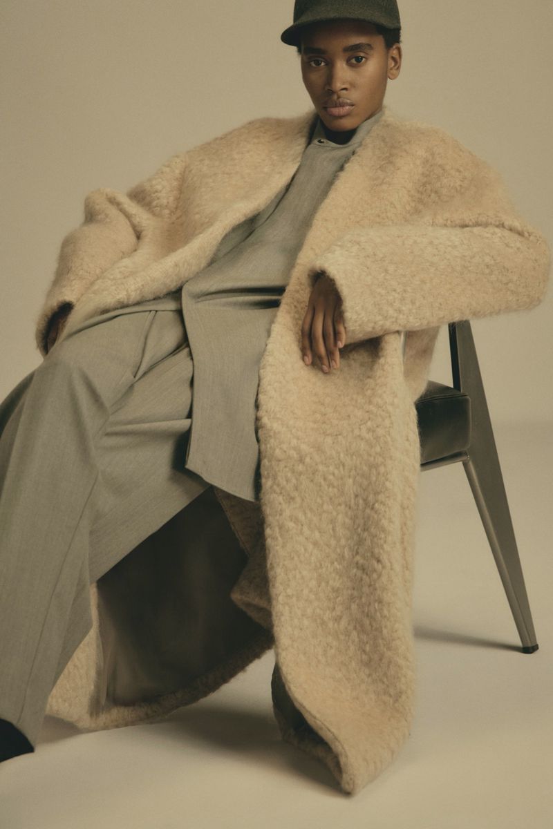 Fashionable woman in oversized beige coat and grey trousers seated in a neutral setting wearing a cap and looking at the camera.