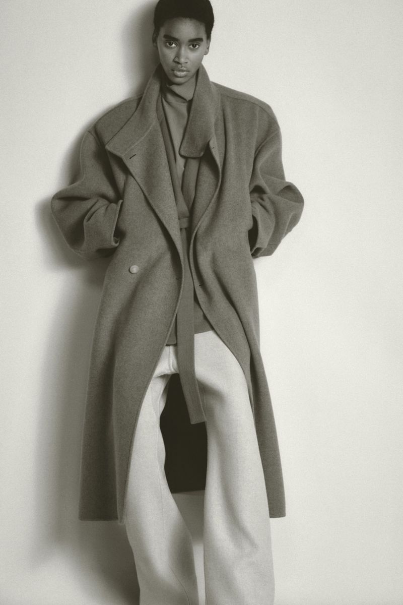 Fashion shot of an African American woman in a warm coat and baggy pants standing indoors in front of a white wall in a studio.