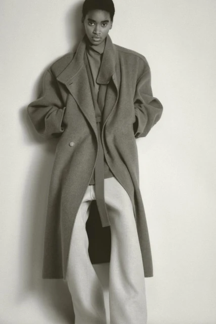 Fashion shot of an African American woman in a warm coat and baggy pants standing indoors in front of a white wall in a studio.