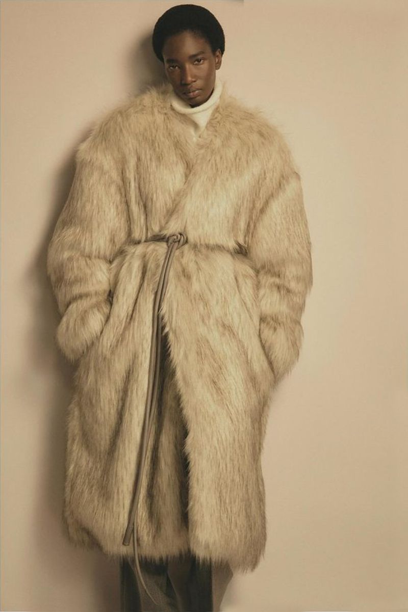 A Black male model poses in a full-length shot wearing a long, beige faux fur coat with a tie belt, black beanie, and white turtleneck sweater.
