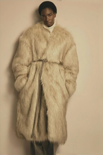 A Black male model poses in a full-length shot wearing a long, beige faux fur coat with a tie belt, black beanie, and white turtleneck sweater.