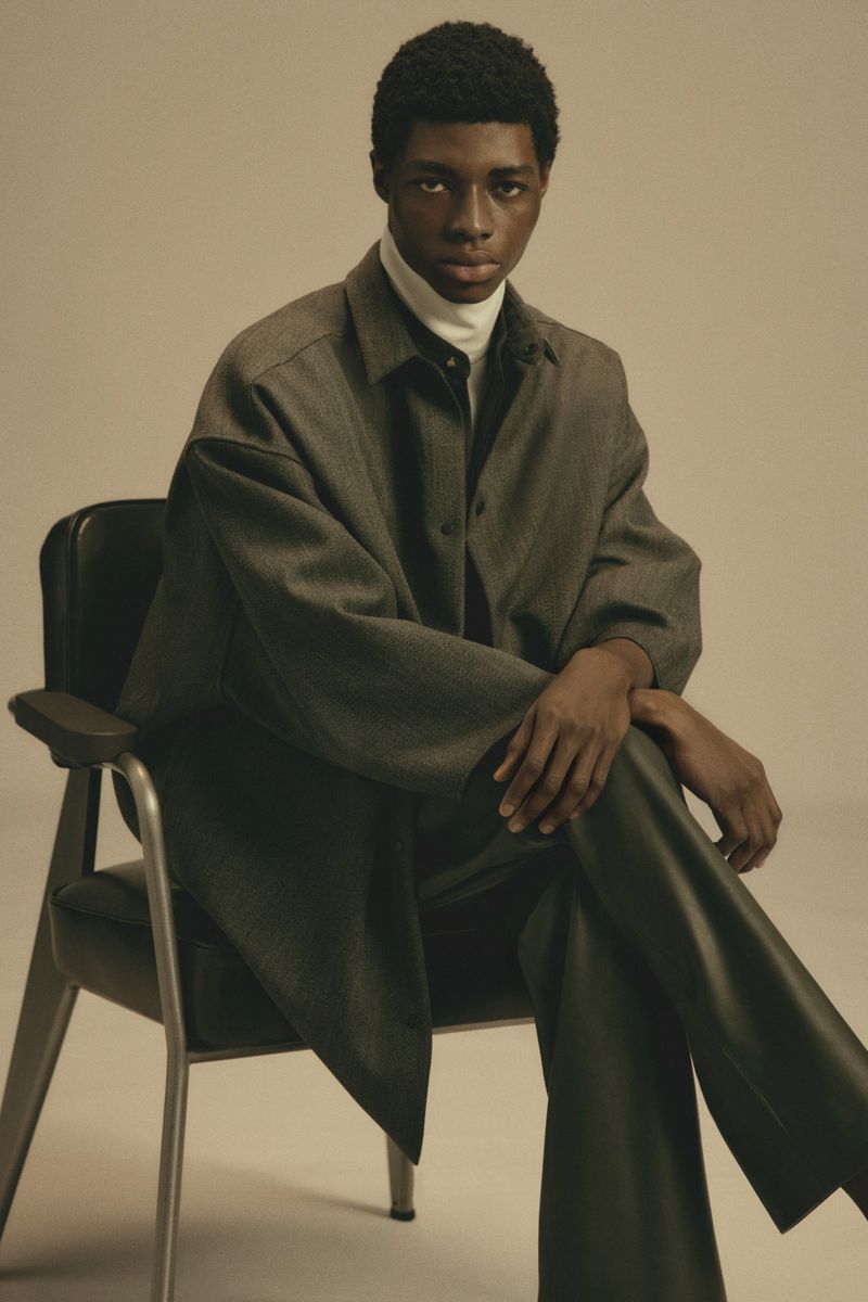 Studio shot of a young Black male model in a grey overshirt and dark pants sitting on a black chair against a beige background.